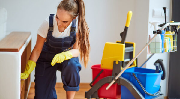 Young staff of cleaning room. Beautiful girl wearing special equipment with yellow rubber gloves, using spray wiping dust off. Careful worker of cleaning company